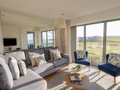 A living room with a gray sofa and blue chairs next to large windows overlooking a grassy area at Newquay Fistral Beach View in Newquay
