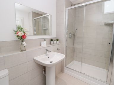 A bathroom with a pedestal sink mirror flowers soap dispenser and a glass shower enclosure at Amble Views in Amble