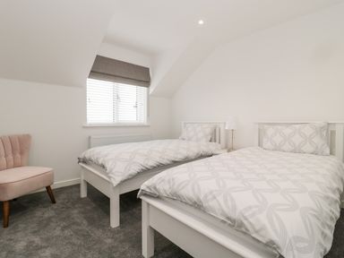 A bedroom with two white single beds a nightstand with a lamp a window with blinds and a pale pink chair at Amble Views in Amble