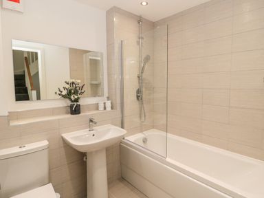 A bathroom with a bathtub and shower, a pedestal sink, a toilet, a large mirror, and flowers on the sink at Amble Views in Amble
