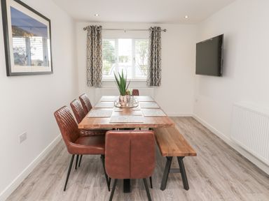 A dining room with a wooden table, brown chairs and a bench, a wall-mounted television and window with patterned curtains at Amble Views in Amble