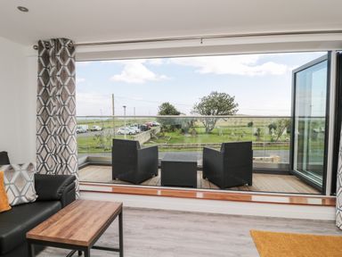 A lounge area with black chairs and a wooden table by large open glass doors leading to a balcony with a view of a grassy area at Amble Views in Amble