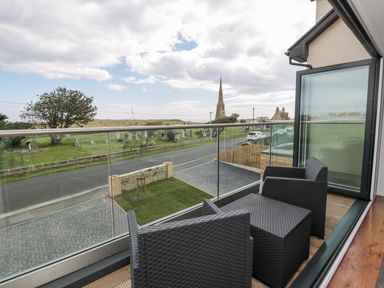 A balcony with two black woven chairs and a small table overlooking a road a graveyard and a church spire at Amble Views in Amble