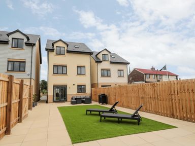 A backyard with artificial grass two black sun loungers patio furniture and beige houses at Amble Views in Amble