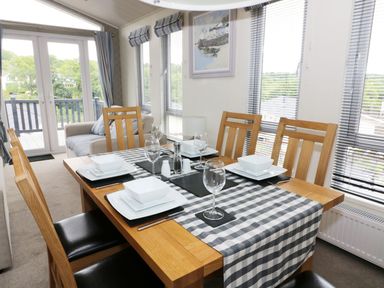 A dining area with a wooden table set with white dishes and wine glasses near windows with blinds and a small sofa at 44 Heritage Park in Stepaside near Kilgetty