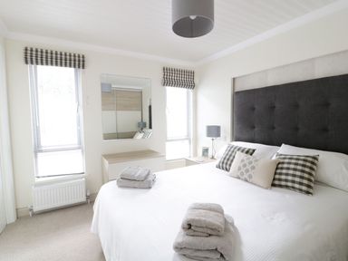 A bedroom with a large bed pillows folded towels bedside table lamp mirror and two windows with blinds at 44 Heritage Park in Stepaside near Kilgetty
