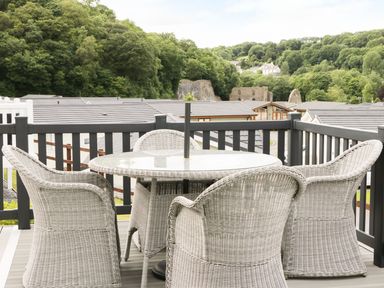 An outdoor deck with four wicker chairs and a round glass table surrounded by a black railing with trees and rooftops in the background at 44 Heritage Park Stepaside near Kilgetty