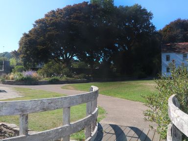 A park area with a wooden fence and pathway surrounded by grass and trees with a white building in the background