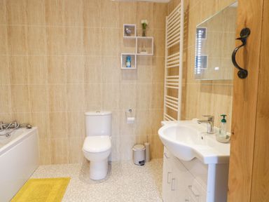 A bathroom with a toilet, sink with cabinet, bathtub, wall shelves, towel rail and a yellow bath mat at Y Stabl in Llanddoged near Llanrwst