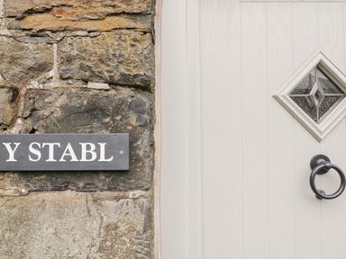 A stone wall with a wooden sign reading Y STABL and a cream door with a diamond glass window and black ring door knocker at Y Stabl in Llanddoged near Llanrwst