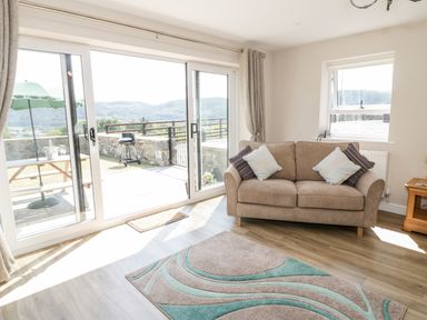 A living room with a brown sofa and cushions near sliding glass doors opening to a patio with a grill and picnic table at Y Stabl in Llanddoged near Llanrwst