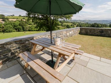 An outdoor patio with a wooden picnic table and green umbrella overlooking a stone wall and green fields at Y Stabl in Llanddoged near Llanrwst