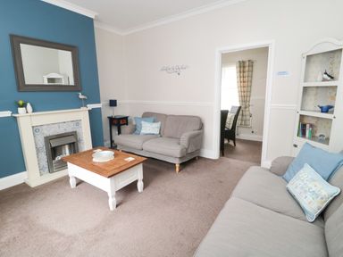 A living room with two sofas a wooden coffee table a fireplace a blue accent wall and a doorway leading to a dining area at Sandy Toes Cottage in Newbiggin-By-The-Sea