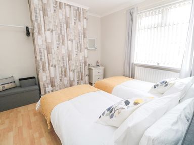 A bedroom with two single beds yellow throws on beds a window with vertical blinds and curtains at Sandy Toes Cottage in Newbiggin-By-The-Sea