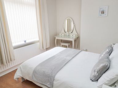 A bedroom with a bed grey cushions and a vanity table by a window at Sandy Toes Cottage in Newbiggin-By-The-Sea