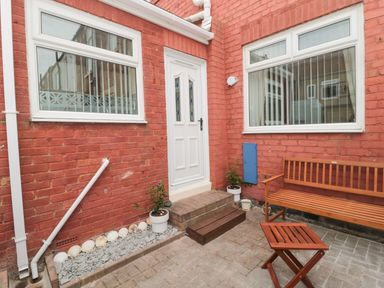 A small outdoor patio area with a white door two windows a wooden bench a small wooden stool and potted plants at Sandy Toes Cottage in Newbiggin-By-The-Sea