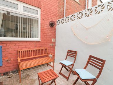 An outdoor patio with wooden bench and chairs with cushions and a decorative net on the wall at Sandy Toes Cottage in Newbiggin-By-The-Sea