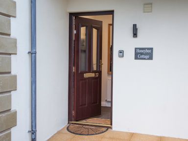 An open brown front door with number 27 and a doormat outside next to a wall with a Honeybee Cottage sign in Berrier near Penruddock