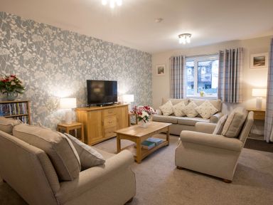 A living room with beige sofas a wooden coffee table a television and floral wallpaper at Honeybee Cottage in Berrier near Penruddock
