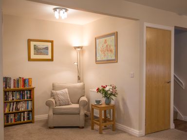 A corner of a room with an armchair a small wooden table with flowers a bookshelf and framed pictures at Honeybee Cottage in Berrier near Penruddock