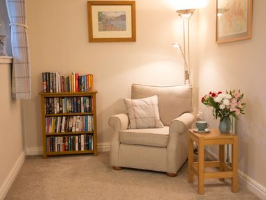 A reading corner with a cushioned armchair a small wooden table with flowers and a cup and a wooden bookshelf with books at Honeybee Cottage in Berrier near Penruddock