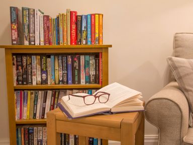 A wooden bookshelf filled with books next to a beige armchair and a small wooden table with an open book and glasses at Honeybee Cottage in Berrier near Penruddock