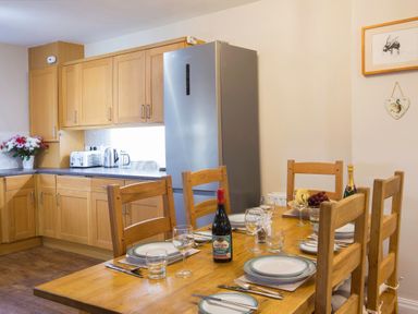 A dining table set with plates glasses cutlery a bottle of wine and fruit in a kitchen with wooden cabinets and a refrigerator at Honeybee Cottage in Berrier near Penruddock