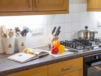 A kitchen countertop with cooking utensils in containers a cookbook a cutting board with bell peppers and a knife a knife block a pepper grinder a pot on a stove at Honeybee Cottage in Berrier near Penruddock