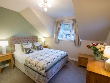 A bedroom with a double bed bedside tables lamps a window with curtains and a wooden dresser with flowers at Honeybee Cottage in Berrier near Penruddock
