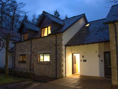 An exterior view of a stone and white wall cottage with lit windows and an open door at Honeybee Cottage Berrier near Penruddock