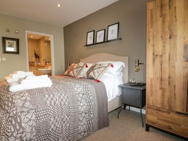 A bedroom with a bed covered in patterned bedding and pillows next to a wooden wardrobe and bedside table with lamp and an open door to a bathroom at The Bolt Hole in Downderry