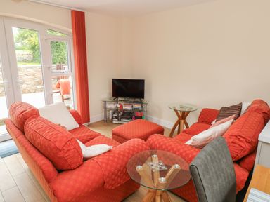 A living room with red sofas a glass coffee table a television on a glass stand and doors leading outside at Nuthatch Barn in Hensol near Pontyclun