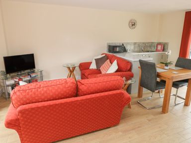 A living room with red sofas a wooden dining table with gray chairs and a small kitchen area at Nuthatch Barn in Hensol near Pontyclun
