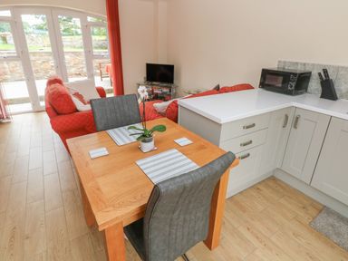 A dining table with two chairs and placemats next to a kitchen counter with a microwave and a knife block and a living area with red sofas and a tv at Nuthatch Barn in Hensol near Pontyclun