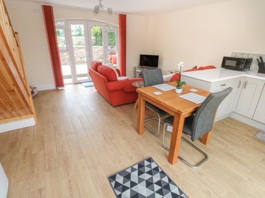 A living area with a wooden dining table, two chairs, a red sofa, a small TV, a microwave, and large windows at Nuthatch Barn in Hensol near Pontyclun