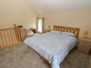 A bedroom with a bed in light blue bedding two wooden bedside tables with yellow lamps a wooden staircase railing a chair by a small window and a wooden cabinet at Nuthatch Barn in Hensol near Pontyclun