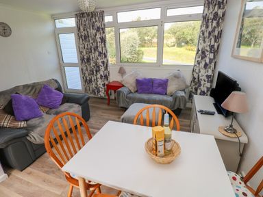 A living room with two sofas purple cushions a white dining table wooden chairs a TV on a cabinet and windows with floral curtains at Y Twll Bollt (The Bolt Hole) in Folkeston Hill Nolton Haven West Wales & Pembrokeshire