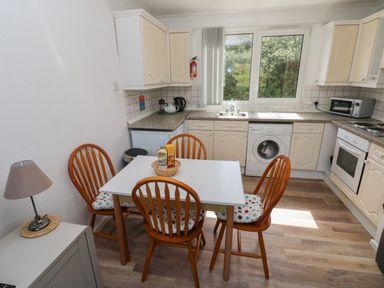 A kitchen with white cabinets wooden chairs a table with a basket and a window at Y Twll Bollt (The Bolt Hole) in Folkeston Hill Nolton Haven West Wales & Pembrokeshire