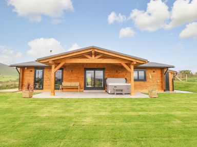 A wooden log cabin with a porch bench and hot tub on a tiled patio surrounded by grass at Bacheiddon Log Cabin Aberhosan near Machynlleth