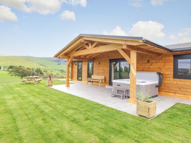 A wooden cabin with a covered patio featuring a bench and hot tub next to a lawn with picnic table at Bacheiddon Log Cabin Aberhosan near Machynlleth