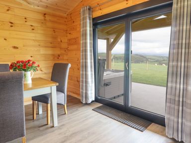 A dining area with a wooden table and gray upholstered chairs next to sliding glass doors showing a patio and grassy hills at Bacheiddon Log Cabin in Aberhosan near Machynlleth