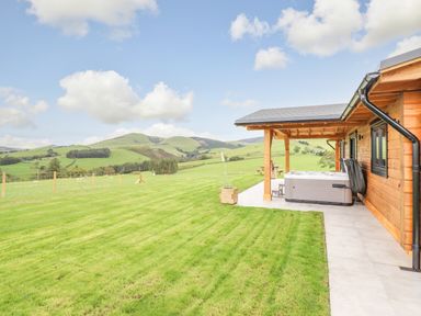 A wooden cabin with patio and hot tub overlooking a fenced grassy field and hills at Bacheiddon Log Cabin in Aberhosan near Machynlleth