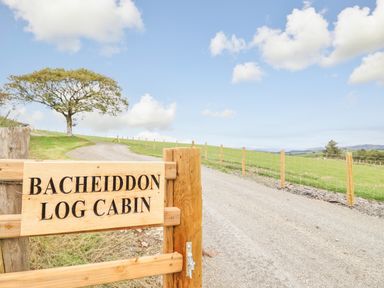 A wooden sign reading Bacheiddon Log Cabin next to a gravel driveway and fenced green fields at Bacheiddon Log Cabin Aberhosan near Machynlleth