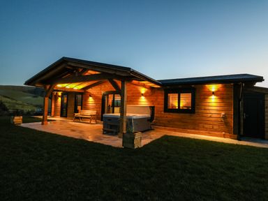 An exterior view of a wooden log cabin with porch lights and a hot tub at Bacheiddon Log Cabin in Aberhosan near Machynlleth