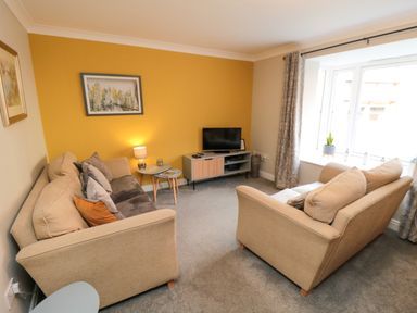 A living room with two beige sofas a yellow accent wall a television on a stand and a window with curtains at Esk Retreat in Whitby