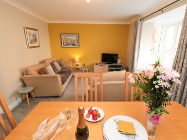 A living room with beige sofas a television and paintings on the walls with a dining table holding a bottle of champagne bread strawberries and flowers at Esk Retreat in Whitby