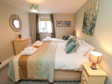 A bedroom with a double bed and a single bed with folded towels on them bedside tables with lamps a chest of drawers with a plant a window with curtains a round mirror and framed pictures on the wall at Esk Retreat in Whitby