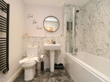 A bathroom with a toilet, pedestal sink, bathtub with shower, round mirror and black towel radiator at Esk Retreat in Whitby
