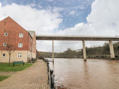 A walkway along a river with a bench and a brick building on one side and a bridge spanning the river at Esk Retreat in Whitby