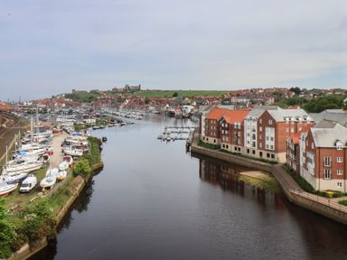 A river with boats docked on the left and buildings on the right with a town and hill in the background at Esk Retreat in Whitby